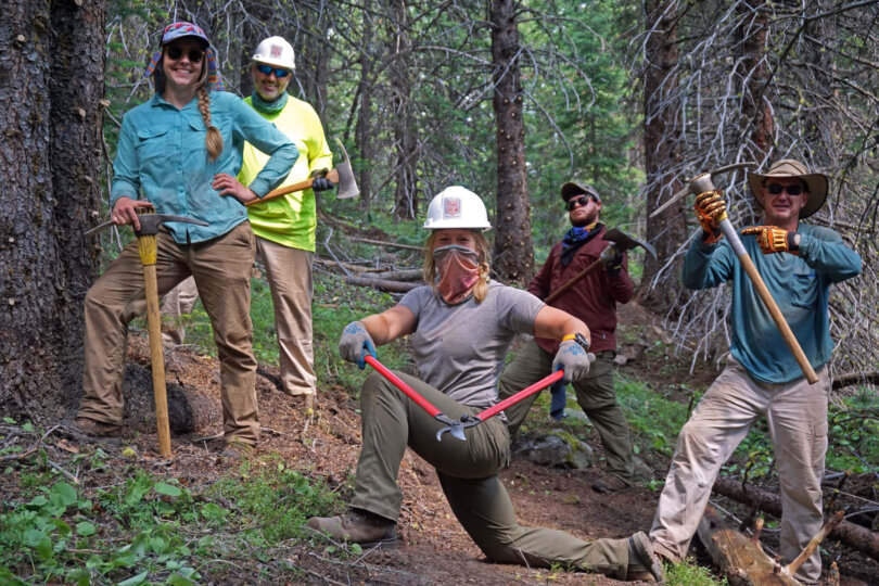 A group of volunteers pose out on the trail holding their trail crew gear