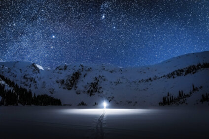 Man standing with light in dark star-filled sky in snowy mountainous landscape.